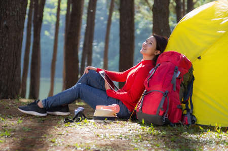 A woman leans against a yellow tent with a red backpack, She is use a tablet and enjoying nature in the midst of a beautiful pine forest beside lake, Pang Oung, Mae Hong Son, Thailand.の写真素材