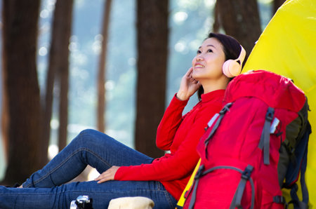 A young girl sits next to a camping tent and a red backpack, She enjoy listening to music from headphones and admiring nature in the middle of forest beside lake, Pang Oung, Mae Hong Son, Thailand.の写真素材