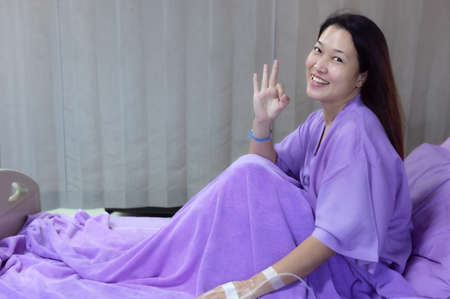 Young Asian female patient is smiling and showing ok gesture. Patient feels happy and comfortable with treatment and therapy on hospital bed in hospital room. Medical healthcare concept.の写真素材