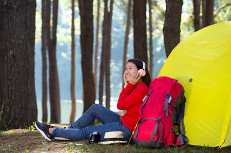 A young girl sits next to a camping tent, She enjoy listening to music from pink headphones and admiring nature in the middle of a pine forest beside the lake, Pang Oung, Mae Hong Son, Thailand.の写真素材