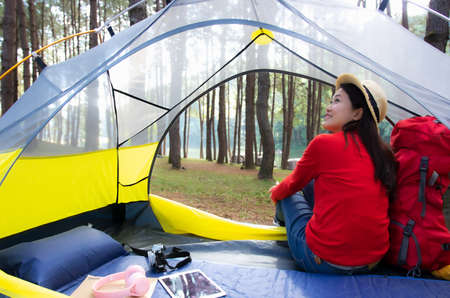 A beautiful woman in a red dress has a camera, an tablet and headphones next to it. She sat in a yellow tent. Admiring the beautiful nature backpacking camping In the middle of a beautiful pine forest beside Pang Oung Lake, Mae Hong Son, Thailandの写真素材