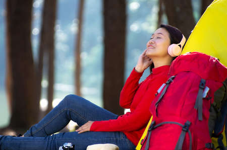 A young girl in a red shirt sits next to a camping tent and a red backpack, There was a camera and a hat next to it.  She enjoy listening to music from pink headphones and admiring nature in the middle of a pine forest beside the lake, Pang Oung, Mae Hong Son, Thailand.の写真素材
