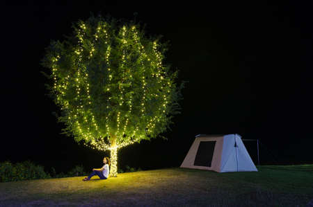 Girl sitting under a tree adorn lights beside tent camping at night. There are beautiful stars in the sky. Chaloem Phrakiat Park, Thung Bua Tong, Mexican sunflower field, Mae Moh, Lampang, Thailand.の写真素材