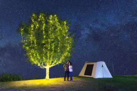 couple under a tree adorn lights beside tent camping at night. There are milky way galaxy stars in the sky. Chaloem Phrakiat Park, Thung Bua Tong, Mexican sunflower field, Mae Moh, Lampang, Thailand.の写真素材