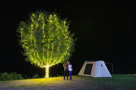 Couple under a tree adorn lights beside tent camping at night. There are beautiful stars in the sky. Chaloem Phrakiat Park, Thung Bua Tong, Mexican sunflower field, Mae Moh, Lampang, Thailand.の写真素材