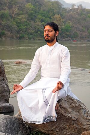 LAXMAN JHULA, INDIA - APRIL 19, 2017: A Hindu swami in white sitting on a rock at the holy river Ganges in Laxman Jhula on the 19th april 2017 in Indiaのeditorial素材