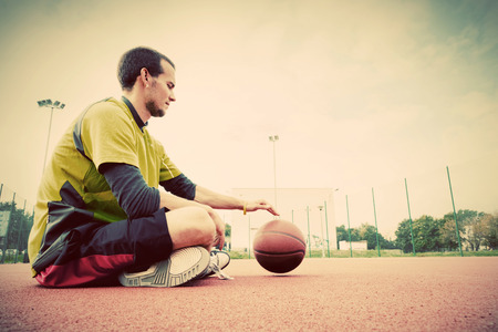 Young man on basketball court. Sitting and dribbling with ball. Streetball, training, activity. Real and authentic, vintage mood.の写真素材