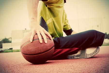 Young man on basketball court. Sitting and dribbling with ball. Streetball, training, activity. Real and authentic, vintage mood.の写真素材