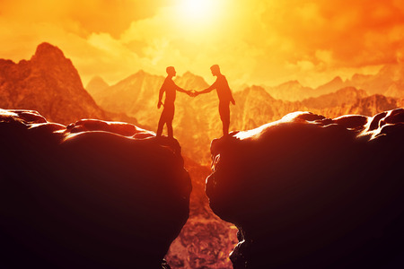 Two men shake hands over precipice between two rocky mountains at sunset. Business, deal, handshake, connection conceptsの写真素材