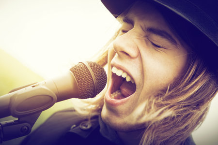 Young man with long hair and hat singing, shouting to microphone. Vintage, music, hipster.