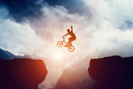 Man jumping on bmx bike over precipice in mountains at sunset. Raising hand showing hello gesture....