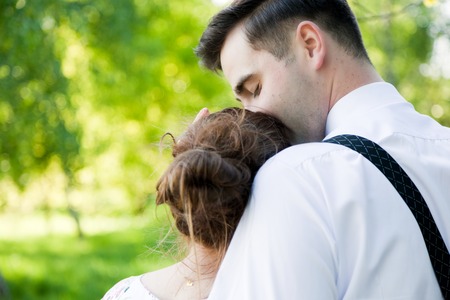 Young handsome man embracing and gently kissing his fiancee in summer green park.  View from the back. Date, fiance with fiancee, couple in love.の写真素材
