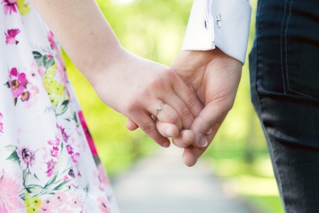 Holding hands close-up. Couple in love dating in summer park. Woman in dress and man wearing elegant shirt. View from the back. Date, fiance with fiancee, hand in hand conceptsの写真素材