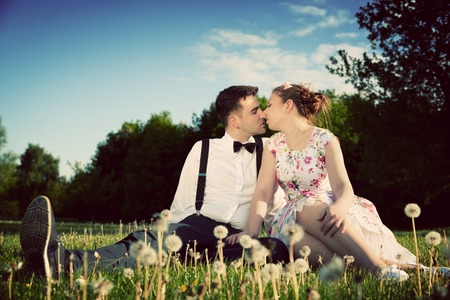 Romantic couple in love about to kiss while sitting on grass in spring park. Vintage date, woman in dress and man wearing suspenders with bow tie.の写真素材
