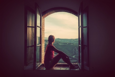 Young woman sitting in an open old window looking on the landscape of Tuscany, Italy. Conceptual romantic, dreaming, hope, travel.の写真素材