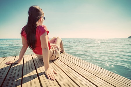 Young woman resting on a wooden jetty looking at the calm sea on sunny summer day. Vintageの写真素材