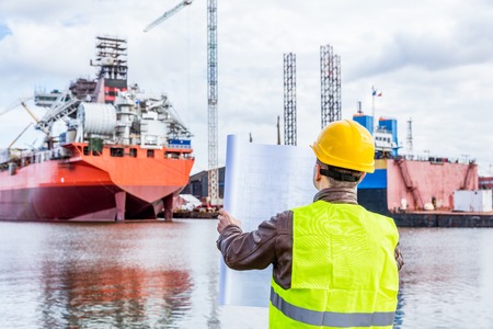 Shipbuilding engineer checking documents and plans of construction at the dockside in a port. Wearing safety helmet and yellow vest, holding folded papers.の写真素材