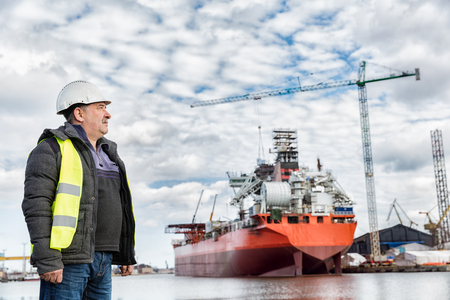 Shipbuilding engineer stands at the dockside in a port. Wearing safety helmet and yellow vest. Shipbuilding industry.の写真素材
