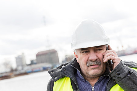 Senior engineer builder talking on the phone at the construction site in a port. Wearing safety helmet and yellow vest.の写真素材