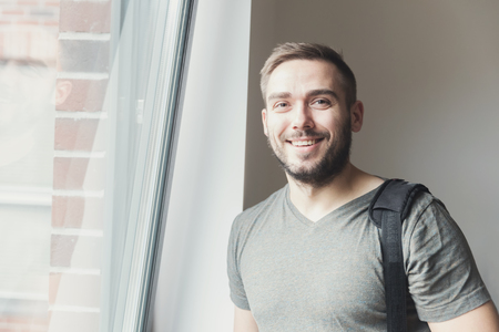 Young man standing by the window with a bag on his shoulder and smiling.の写真素材