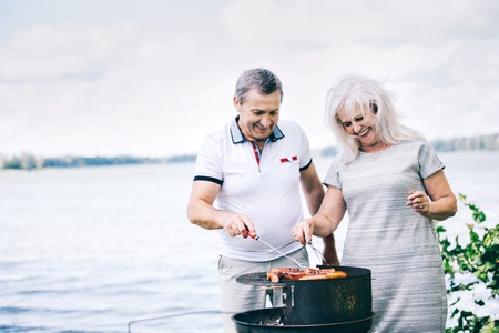 Cheerful senior couple preparing sausages on the barbeque. Summertime activities.の写真素材