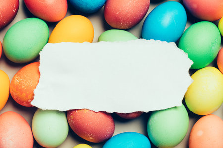 Piece of plain white paper laying on the top of colorful eggs. Easter. Copyspace.