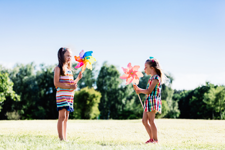 Two little girls blowing upon colorful pinwheels in the park. Friends playing outdoors.の写真素材