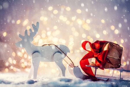 Toy sleigh with a Christmas gift pulled by a wooden reindeer in snow. Glitter lights background.の写真素材