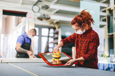 Man and woman in face masks working on glass pane in glazier workshop. Industry and work during coronavirus Covid-19の写真素材
