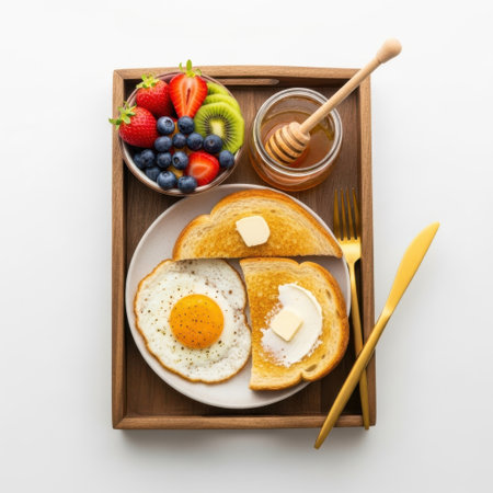 A wooden tray holds a variety of breakfast items. There is a fried egg with a runny yolk on a slice of toast, accompanied by another slice of toast with butter. Next to the toast, there is a small bowl filled with a colorful assortment of fresh fruits including strawberries, blueberries, kiwi, and a few other fruits. A jar of honey with a wooden dipper is also present on the tray, along with a fork and a knife. The arrangement is neatly organized, suggesting a wholesome and nutritious morning meal.の素材