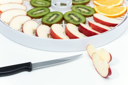 Kitchen knife and cut apple. Behind is a tray of dehydrator with slices of kiwi and apples.の写真素材