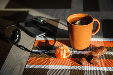A cup of tea, headphones, smartphone, tangerine and a piece of chocolate are on the kitchen table.の写真素材