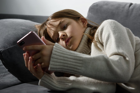 The young girl is looking at the screen of a smartphone leaning on a sofa pillow.