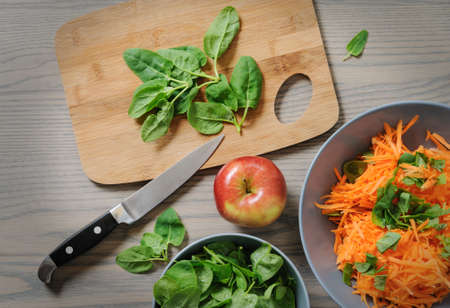 Fresh spinach is on a cutting board. There is an apple, a kitchen knife, a bowl with grated carrots and a bowl with spinach on a wooden table.の写真素材