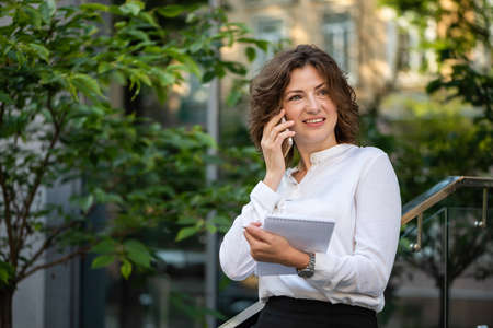 Beautiful business woman is talking on a smartphone. She is holding a notebook.の写真素材