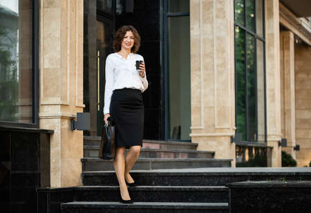 Beautiful business woman with a cup of coffee is coming out of a business building.の写真素材