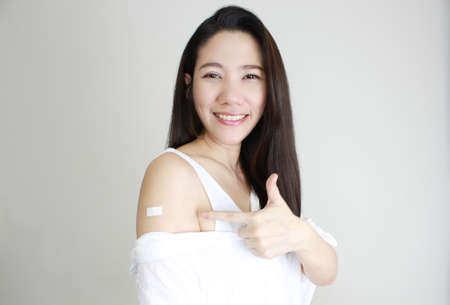 Asian happy smile beautiful women wear white shirt receive covid-19 anti virus vaccine and indicate finger on white bandage with white backgroundの写真素材