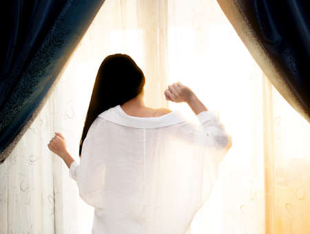Asian woman wake up and standing at window and looking out and stretching in sunlight in the morning.の写真素材