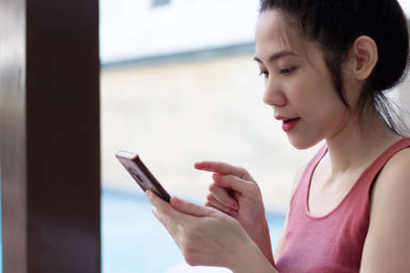 Asian Beautiful women wear pink sleeveless t-shirt holding smartphone are on social media or chatting with family or shopping online on her holiday and weekend in leisure day at her homeの写真素材
