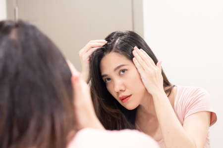 Asian Beautiful woman are looking herself in mirror and use hand touch her hair and looking for  grey hair or hair loss with a worry face.の写真素材