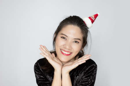 Asian beautiful woman wear small red merry X'mas hat and raise palm hand under chin with smiling and a happiness to celebrating Christmas and happy new yearの写真素材