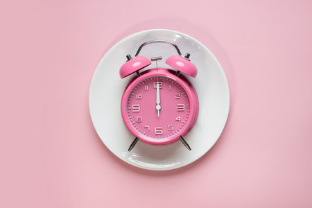 Flat lay of pink alarm clock on white plate on pink background.  Ketogenic dieting, weight loss,...