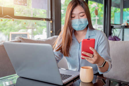 Thai female office worker Put on a mask I am using a red smartphone and working on my laptop in a coffee shop during the day. She was wearing a blue shirt and sipping coffee.の写真素材