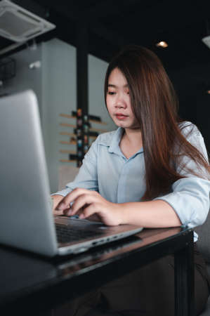 A female Asian office worker of Thai descent is typing and working with notebooks in a coffee shop during the day. She was wearing a blue shirtの写真素材