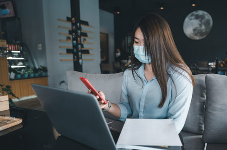 Asian woman wearing a mask uses a smartphone in a coffee shop.