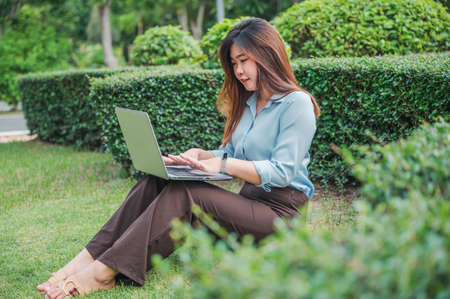 Asian female office worker using laptop or computer  notebook in the park She wears a blue shirt.の写真素材