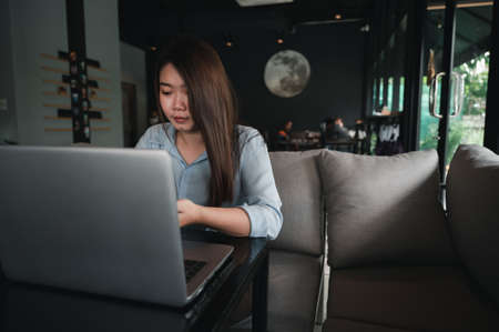 A female Asian office worker of Thai descent is typing and working with notebooks in a coffee shop during the day. She was wearing a blue shirtの写真素材