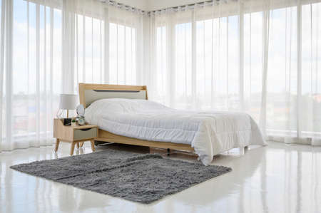 The bedroom is decorated in white tones. There are sun blinds and the sun shines lightly on the window. There is a lamp and an alarm clock at the head of the bed. Modern bedroom in bright white.の写真素材