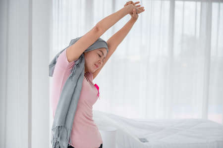 Young asian women wearing hijab Open the curtains in the bedroom to get the morning sunshine. Attaching a pink ribbon represents recovery from a breast cancer patient. Breast cancer conceptの写真素材