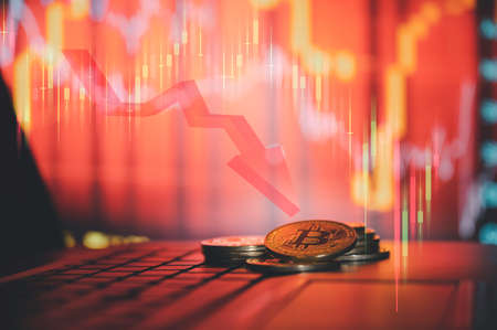 Bitcoins are falling heavily. Crypto currency is placed on the keyboard of the laptop with a backdrop of the falling stock graph.の写真素材
